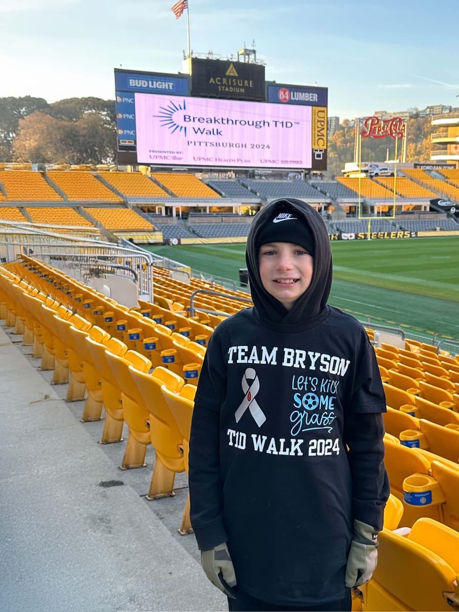 Bryson in the stands at Acrisure Stadium wearing his black Team Bryson walk shirt, with the Breakthrough T1D Walk scoreboard behind him