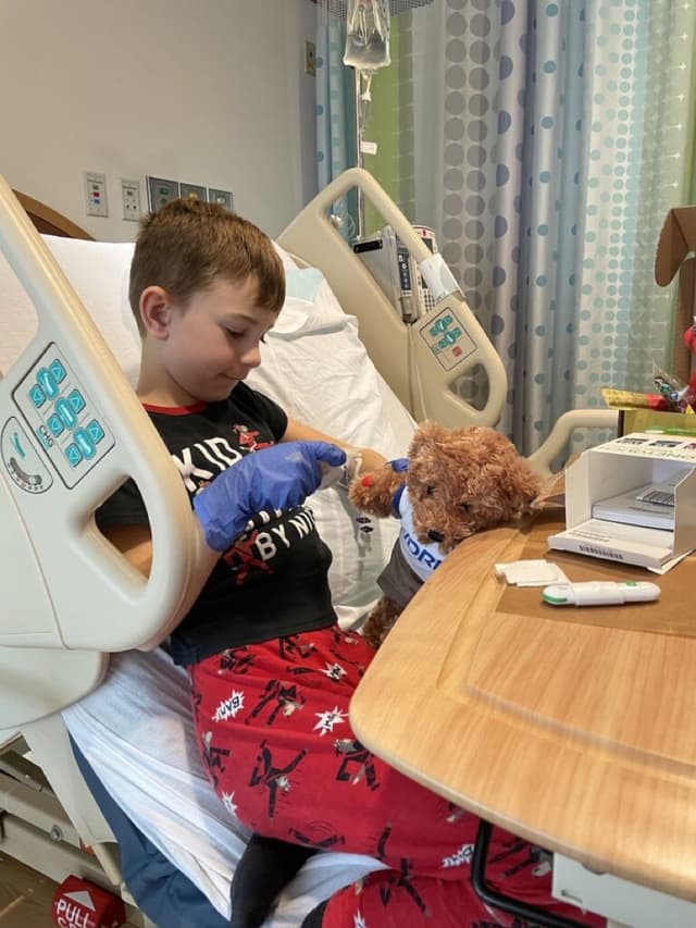 Bryson in a hospital bed wearing a blue glove, practising an injection on Rufus, the Breakthrough T1D bear